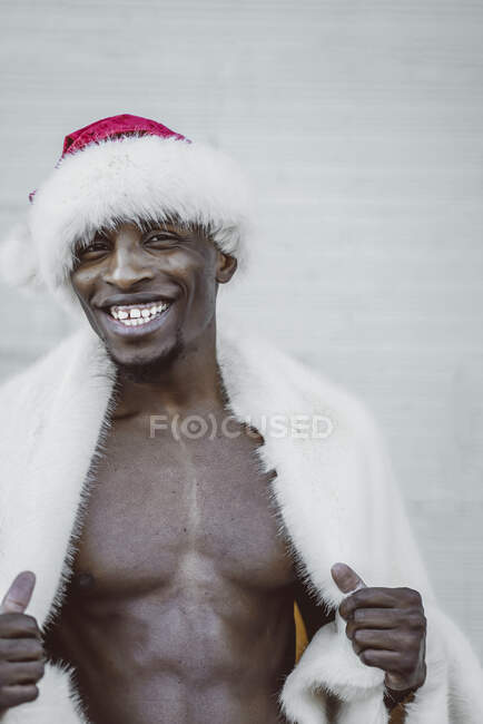 Handsome African American man in fur coat and Santa Claus hat smiling and looking at camera while standing near white wall — Stock Photo