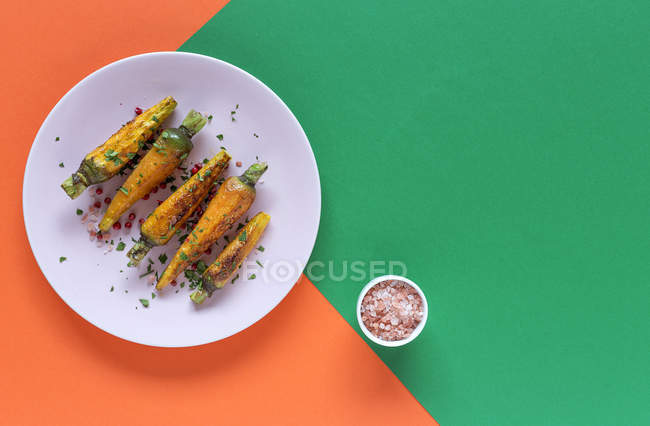 Gesunde geröstete Karotten mit Kräutern und Gewürzen auf Teller auf grünem und orangefarbenem Hintergrund mit Salz — Stockfoto