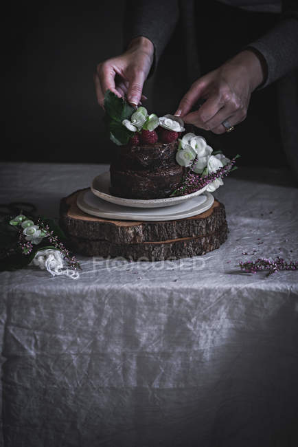 Frauenhände dekorieren Schokoladenkuchen mit Himbeeren und Blumen auf Teller auf Holzständer — Stockfoto