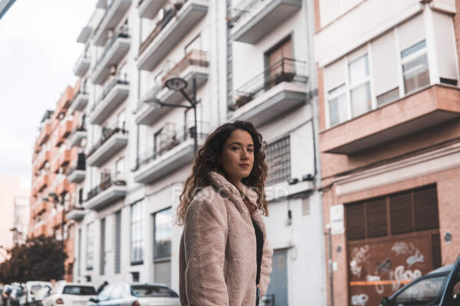 Serious young woman with curly hair looking at camera while standing on city street — Stock Photo
