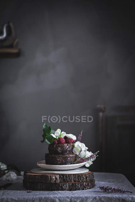 Шоколадный торт, украшенный малиной и цветами, подается на тарелке на деревянном стенде — стоковое фото