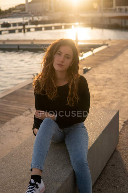 Позитивна молода жінка дивиться на камеру, сидячи біля води на міській набережній під час заходу сонця — стокове фото