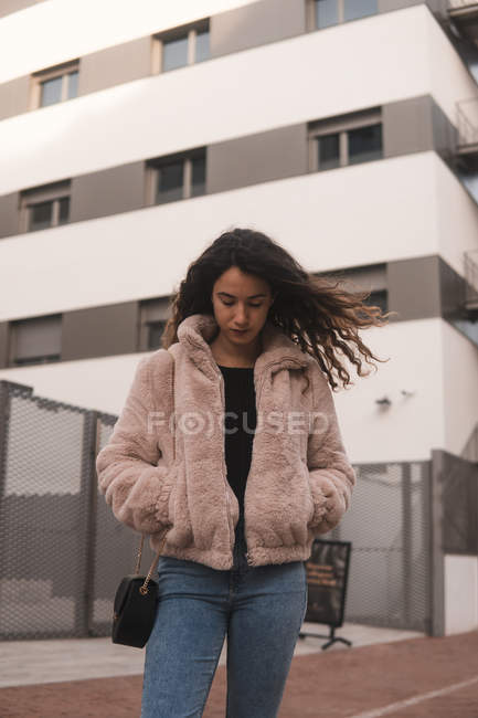 Portrait of young woman with curly hair and hands in pockets standing near modern building on city street — Stock Photo