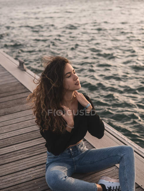 Charming young female with eyes closed sitting on wooden pier near water — Stock Photo