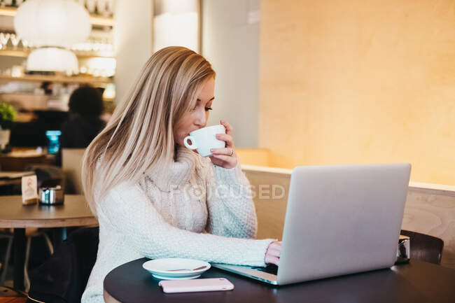 Señora usando el ordenador portátil en la mesa con taza de bebida y teléfono inteligente - foto de stock