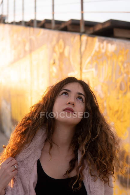 Young woman in trendy outfit touching curly hair while standing near stadium stand on sunny day — Stock Photo
