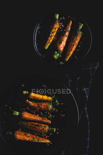 Zanahorias asadas saludables con hierbas y especias en platos negros sobre fondo oscuro - foto de stock