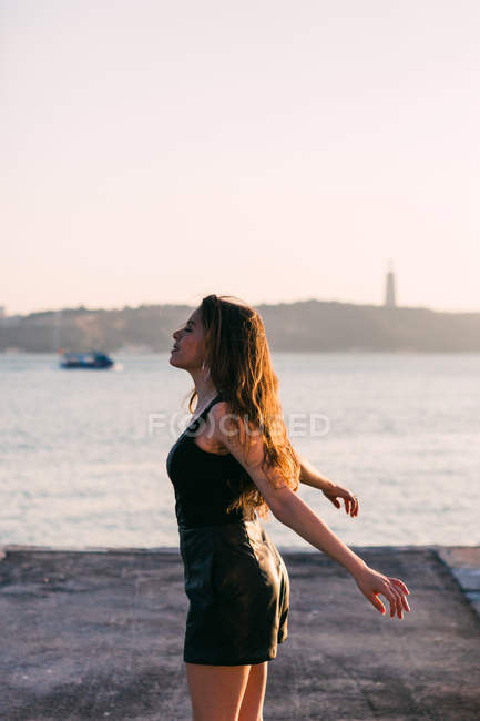 Весела жінка в чорному одязі танцює на набережній біля поверхні води на заході сонця — стокове фото