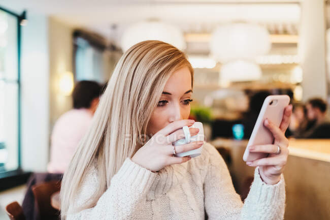 Женщина с помощью смартфона за столом с чашкой напитка — стоковое фото