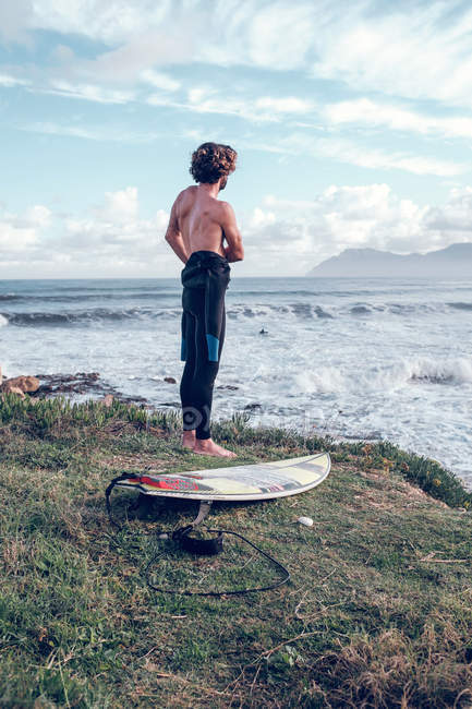 Young man with surfboard putting on wetsuit near ocean — Stock Photo