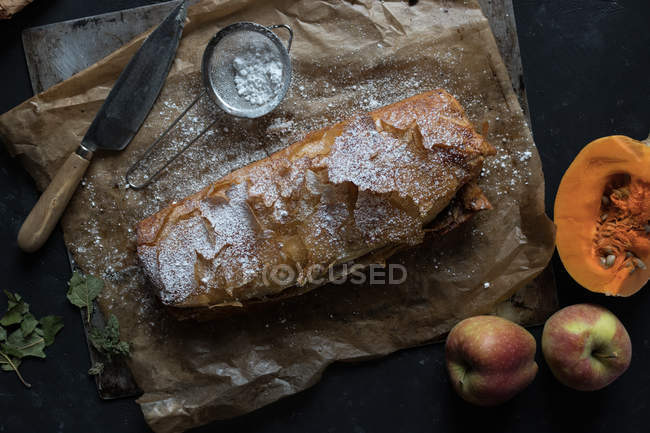 Calabaza casera y strudel de manzana en pergamino con ingredientes sobre fondo oscuro - foto de stock