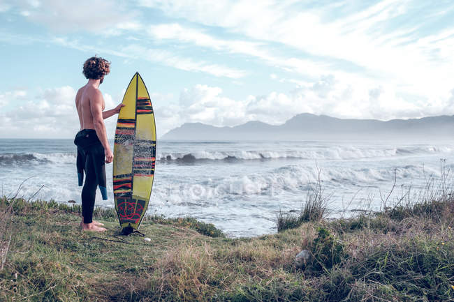Guy debout avec planche de surf lumineuse sur la côte près de l'océan avec planche de surf — Photo de stock