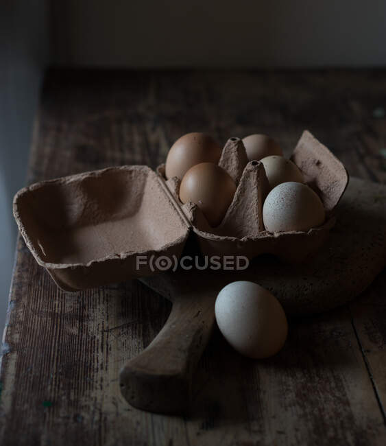 Carton with fresh chicken eggs lying on cutting board on wooden table in dark room — Stock Photo