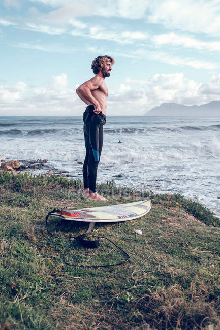 Young man with surfboard putting on wetsuit near ocean — Stock Photo
