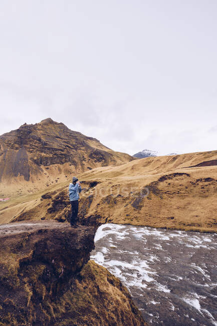 Парень с боковым видом, стоящий на камне возле течения реки между коричневыми горами в Исландии — стоковое фото