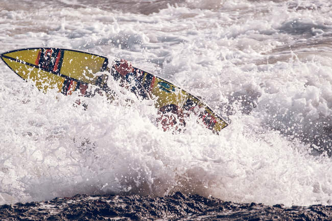 Tabla de surf brillante en olas y salpicaduras de mar cerca de la costa rocosa - foto de stock