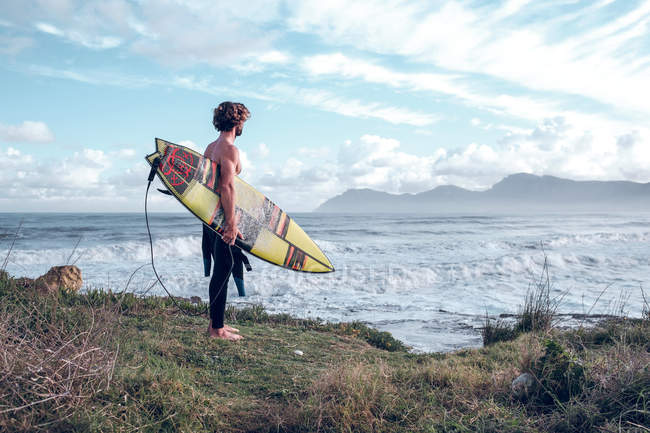Cara de pé com prancha brilhante na costa perto do oceano com prancha de surf — Fotografia de Stock