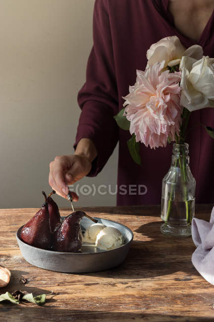 Cropped of woman tasting with spoon bowl of vanilla ice cream and mulled pears in red wine on table with flowers in vase — Stock Photo