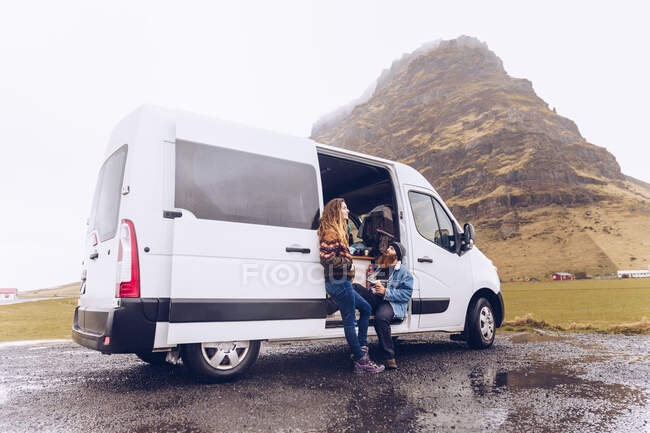 Beard guy and attractive lady in warm wear with mug looking away near opened van door near rock hill in Iceland — Stock Photo