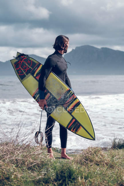 Молодий чоловік у гідрокостюмі з дошкою для серфінгу, що стоїть на морському узбережжі і дивиться на погляд — стокове фото