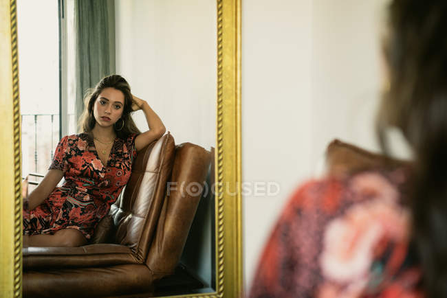Riflessione della donna bruna attraente appassionata in abito con motivo floreale e seduta a testa in testa sul divano in camera — Foto stock