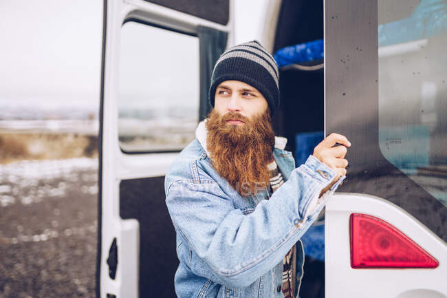 Vista laterale ragazzo barba rossa in giacca di jeans e cappello guardando lontano vicino aperto? van porte in Islanda — Foto stock