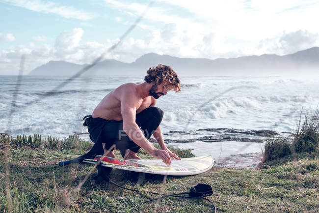 Young man cleaning surfboard on ocean coast — Stock Photo