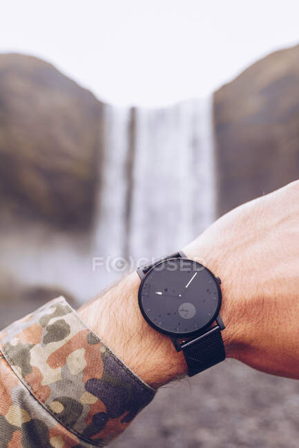 Mano recortada del tipo que muestra relojes negros cerca de la cascada de agua en Islandia sobre un fondo borroso - foto de stock
