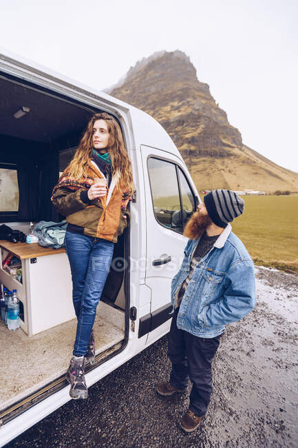 Barba chico y atractiva dama en ropa de abrigo con taza mirando hacia otro lado cerca de puerta abierta furgoneta cerca de roca colina en Islandia - foto de stock