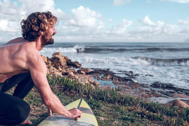 Молодой человек чистит доску для серфинга на берегу океана и смотрит на вид — стоковое фото