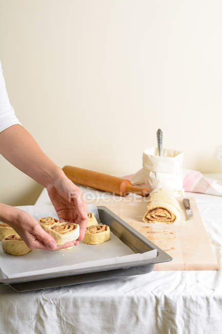 Cortado de manos de mujer poniendo bollos remolinados en bandeja para hornear en la cocina . - foto de stock