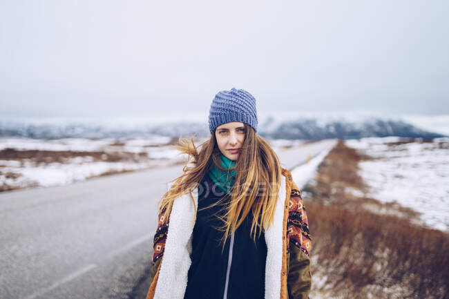 Молода приваблива жінка в гірськолижній куртці і капелюсі дивиться на камеру на дорозі між дикими землями в снігу в Ісландії. — стокове фото