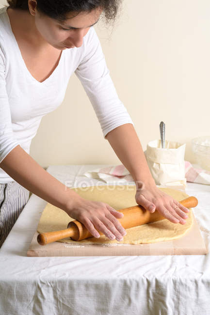 Jolie femelle utilisant un rouleau en bois pour rouler la pâte fraîche dans une cuisine confortable — Photo de stock