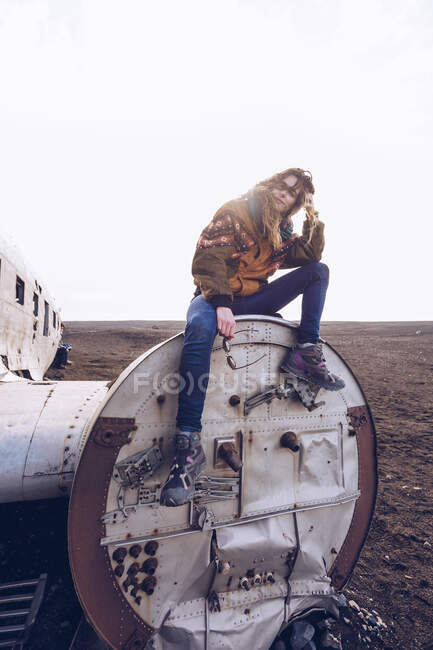 Девушка в теплой одежде сидит на сломанном самолете между темными землями в Исландии — стоковое фото