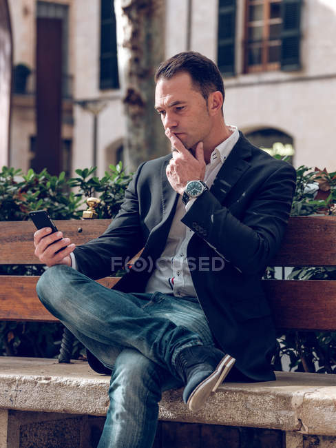Bello uomo adulto in giacca elegante che tiene lo smartphone e distoglie lo sguardo mentre si siede sulla panchina in strada — Foto stock