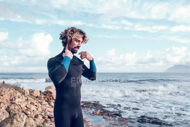 Joven barbudo poniéndose traje de neopreno cerca del océano - foto de stock