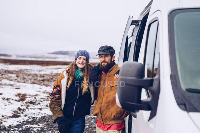 Barba chico y atractiva dama alegre en ropa de abrigo mirando la cámara cerca de la puerta abierta furgoneta entre el campo en Islandia - foto de stock