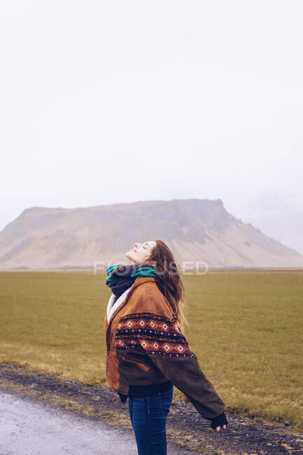 Вид сзади молодая привлекательная леди в? пальто глядя на камеру на дороге между дикими землями с каменными холмами в Исландии — стоковое фото