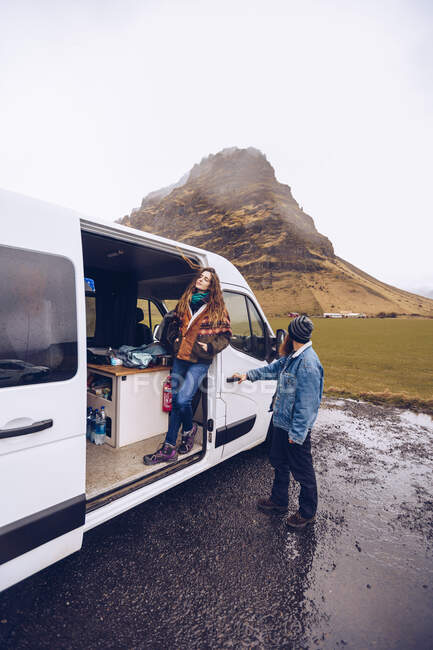 Barba chico y atractiva dama en ropa de abrigo mirando hacia otro lado cerca de la puerta abierta furgoneta cerca de roca colina en Islandia - foto de stock