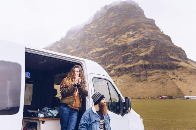 Хлопець і приваблива жінка в теплому одязі з кухлем, що дивиться далеко відкритими дверима фургона біля скелястого пагорба в Ісландії — стокове фото