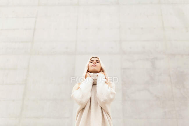 Cute young female in elegant warm sweater looking up while standing near white wall on city street — Stock Photo