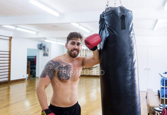 Портрет усміхненого молодого боксера, що спирається на мішок для ударів у спортзалі — стокове фото