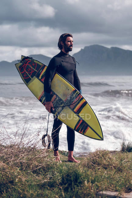 Jeune homme en combinaison avec planche de surf marchant sur la côte — Photo de stock