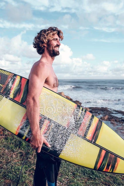 Sonriente tipo de pie con tabla de surf brillante en la costa cerca del océano con tabla de surf - foto de stock