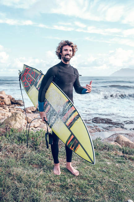 Portrait of young man in wetsuit with surfboard standing near sea and showing thumb up — Stock Photo