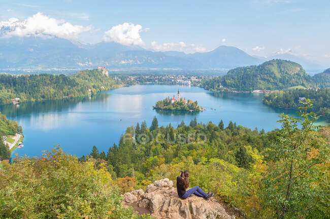 Back view of lady with camera sitting on rock and shooting landscape of lake between forest and town near mountains in Slovenia and Croatia - foto de stock