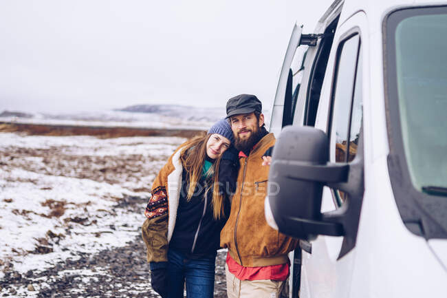Barba chico y atractiva dama alegre en ropa de abrigo mirando la cámara cerca de la puerta abierta furgoneta entre el campo en Islandia - foto de stock
