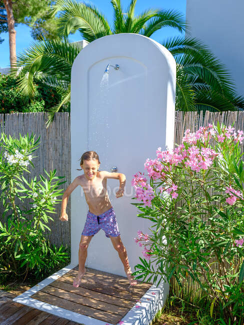 Garoto bonito em shorts de natação sorrindo e de pé sob a água do chuveiro no dia ensolarado em maravilhoso resort — Fotografia de Stock