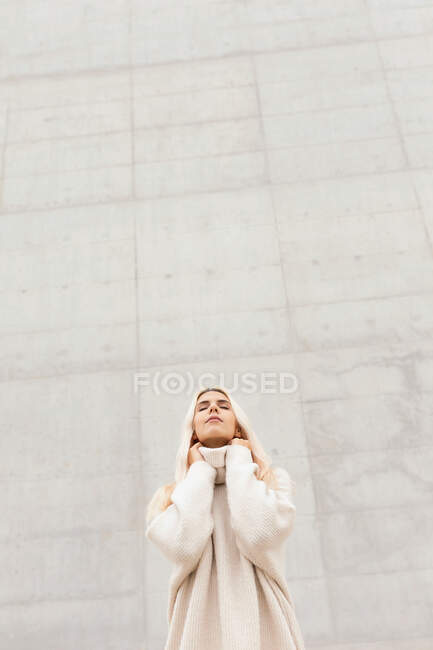 Cute young female in elegant warm sweater looking up while standing near white wall on city street — Stock Photo