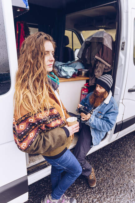 Beard guy and attractive lady in warm wear with mug looking away near opened van door near rock hill in Iceland — Stock Photo